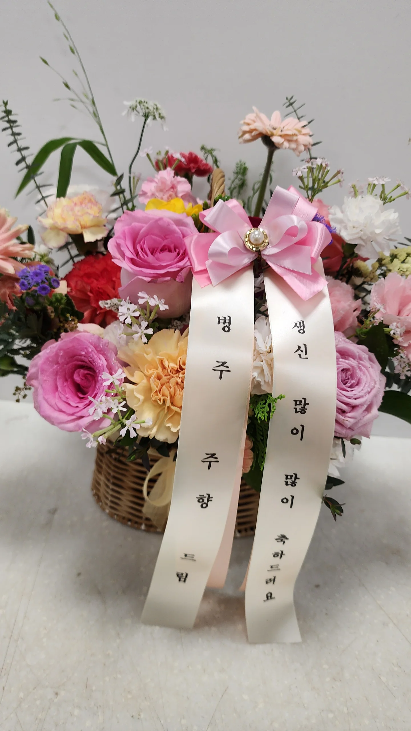 분홍 장미와 흰 꽃이 어우러진 꽃바구니에 긴 리본 장식이 달린 상품이다.