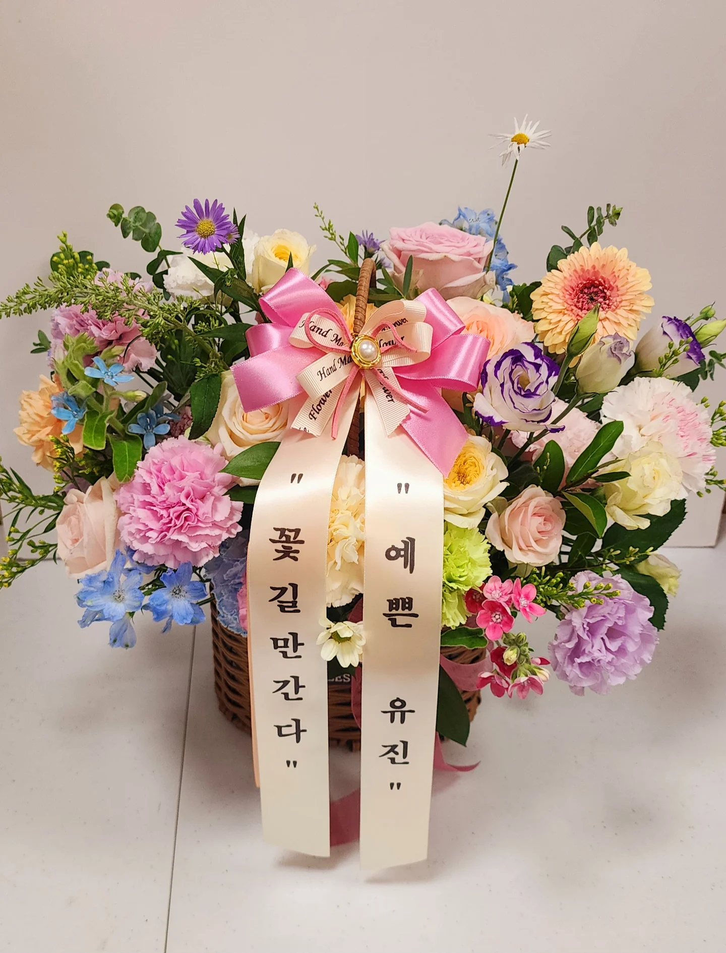 핑크와 퍼플, 옐로우 꽃이 풍성하게 어우러진 바구니형 꽃 상품에 큰 리본 장식이 달려 있다.