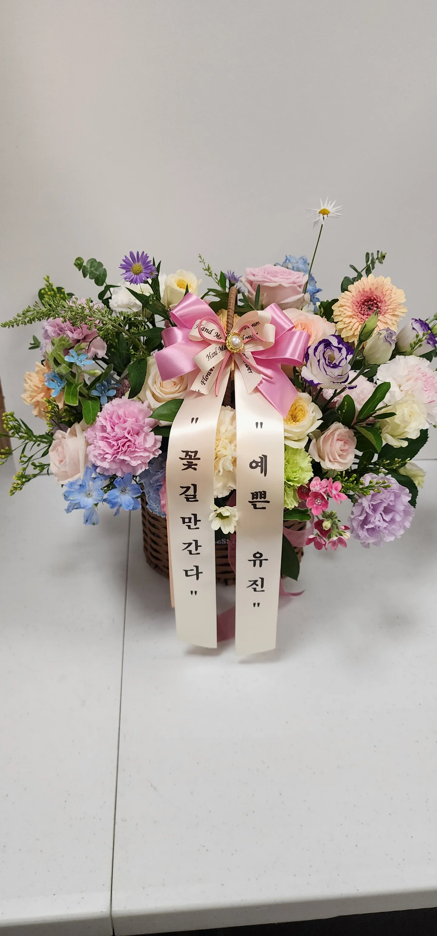 핑크, 화이트, 퍼플 꽃이 풍성하게 꽂힌 꽃바구니에 큰 리본과 축하 문구 리본이 달려 있다.