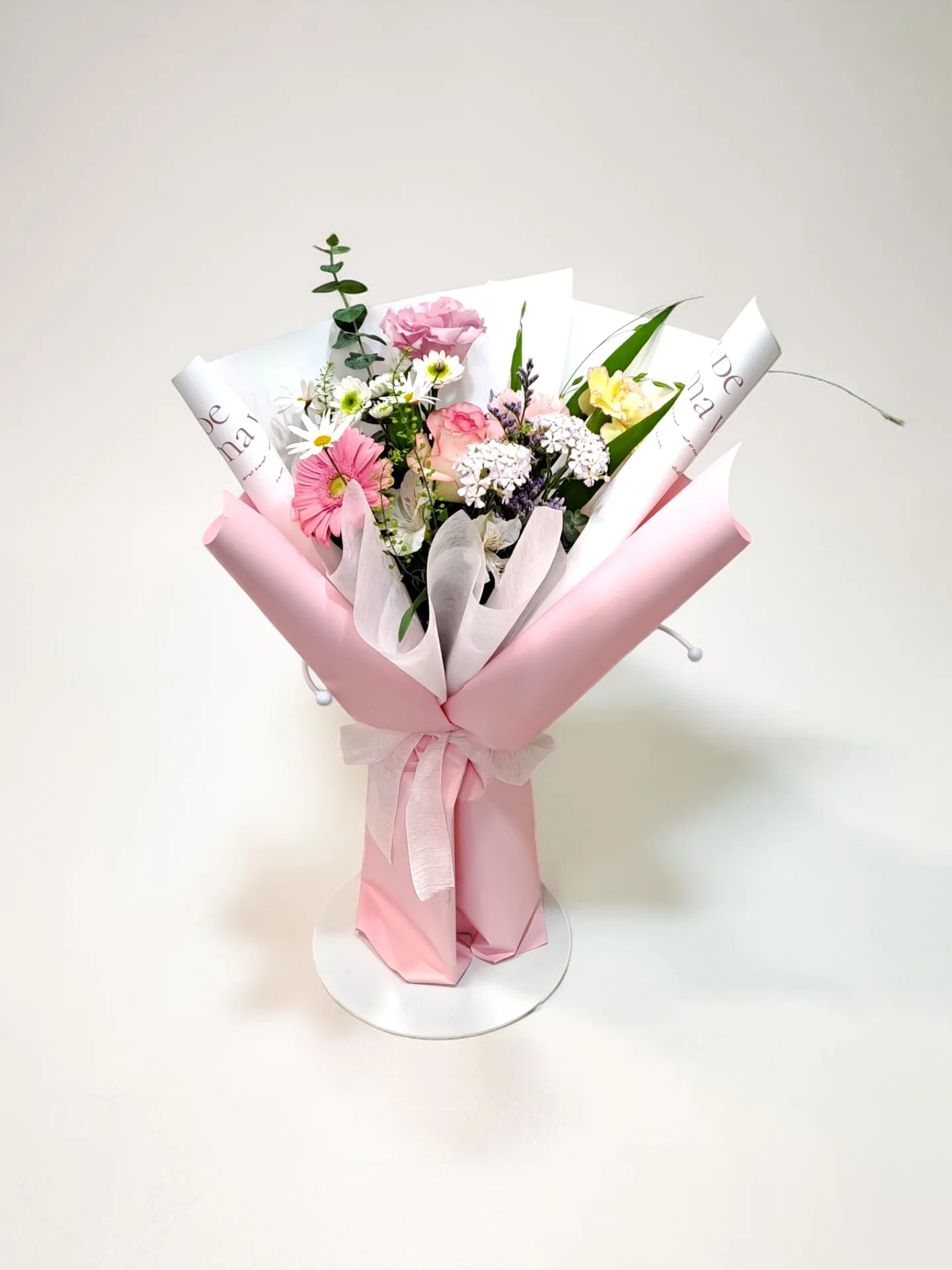 핑크 거베라와 카네이션, 작은 흰색 꽃들이 섞인 꽃다발이 연핑크 포장지로 감싸여 있다.